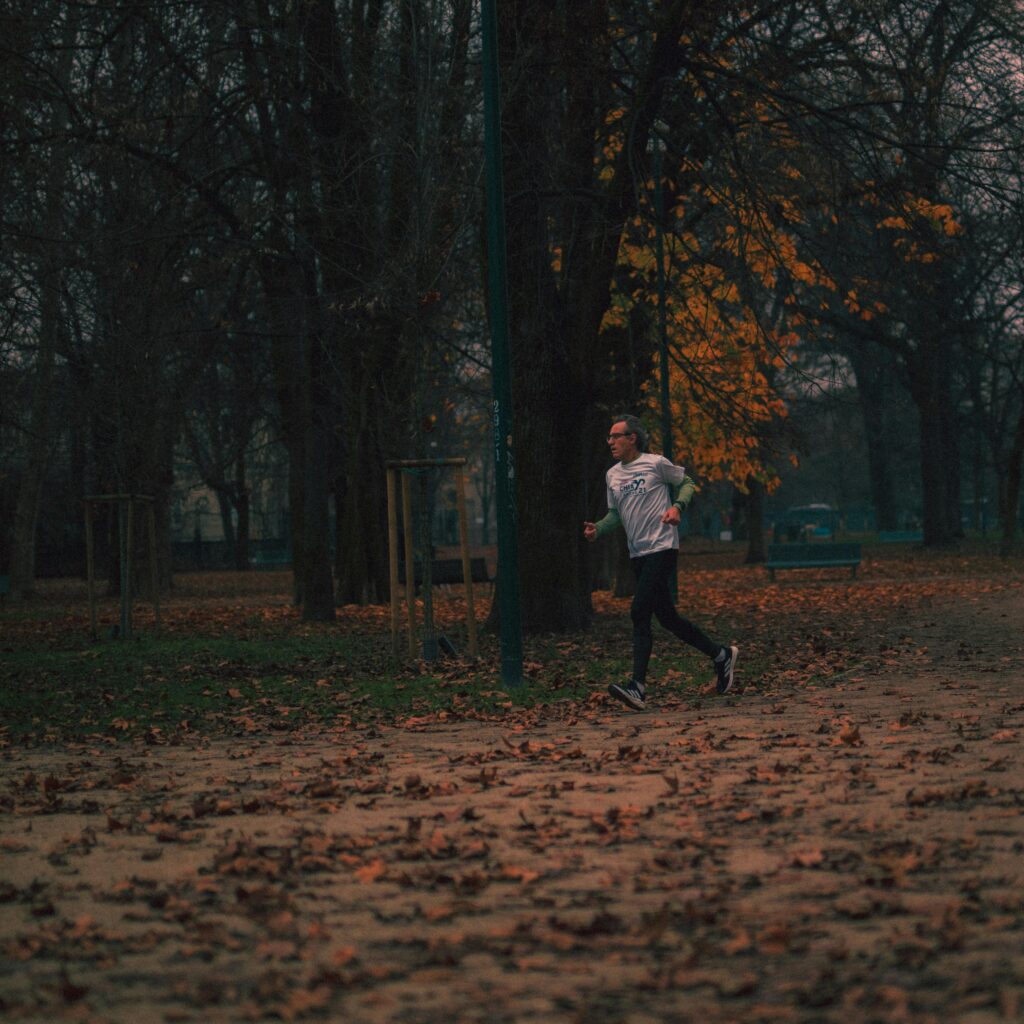A man jogging in a park during fall, surrounded by fallen leaves and bare trees.